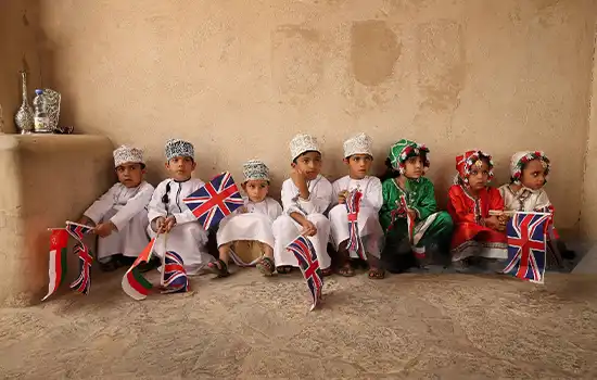 An image of a group of Omani children in traditional attire, holding the flags of Oman and the United Kingdom.