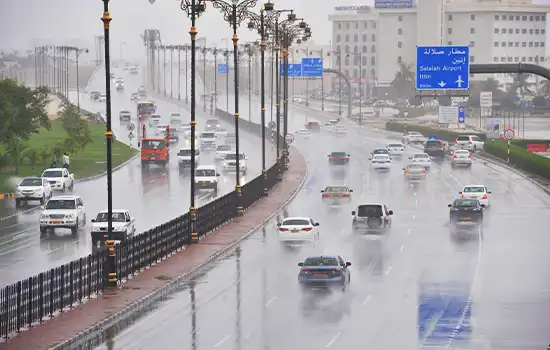A rain-soaked street in Oman with light traffic and road signs pointing to Salalah Airport