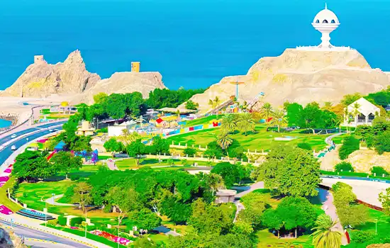 An aerial view of a lush green park in Muscat, featuring landscaped gardens, walking paths, and the iconic Al Riyam Incense Burner structure atop a hill with the Arabian Sea in the background.